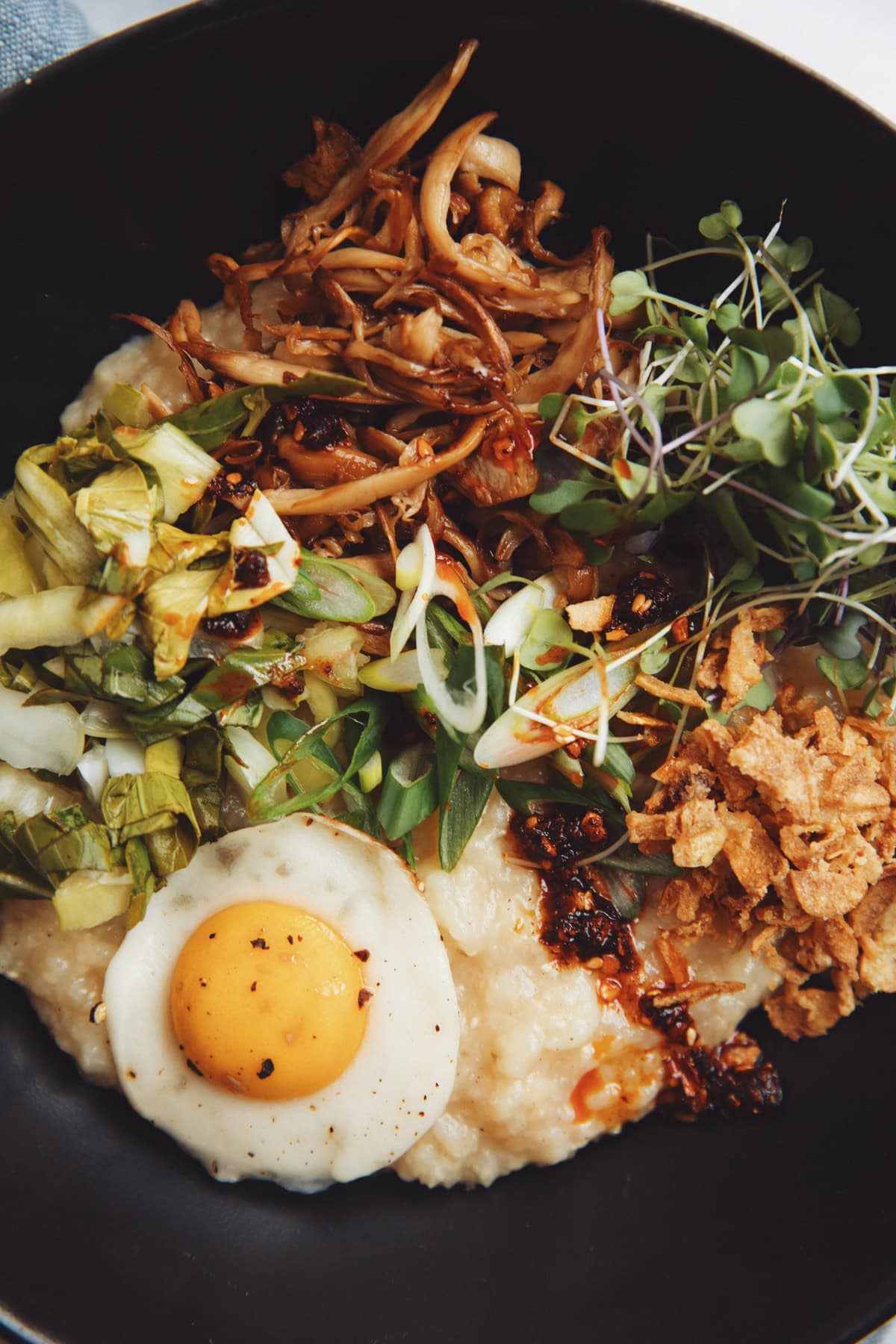 close up of vegan breakfast congee bowl topped with shredded king oyster mushrooms, pickled bok choy, crispy fried onions, micro greens, and scallions